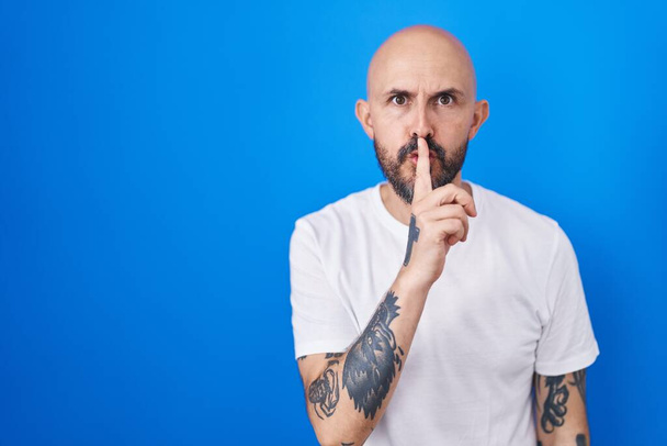 Spanyol férfi tetoválásokkal a kék háttér fölött, és azt kéri, hogy maradjon csendben, ujjal a száján. csend és titkos koncepció.  - Fotó, kép