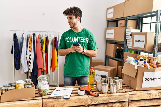 Jonge Spaanse man in vrijwilligersuniform in gesprek op de smartphone in het liefdadigheidscentrum - Foto, afbeelding
