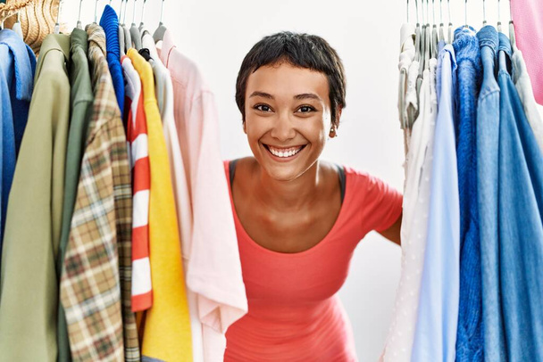 Νεαρή Ισπανίδα πελάτισσα χαμογελά με αυτοπεποίθηση εμφανιζόμενη μέσα από ρούχα στο κατάστημα ρούχων - Φωτογραφία, εικόνα