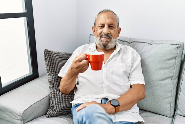 Idősebb ősz hajú férfi, aki magabiztosan mosolyog, miközben otthon kávézik. - Fotó, kép