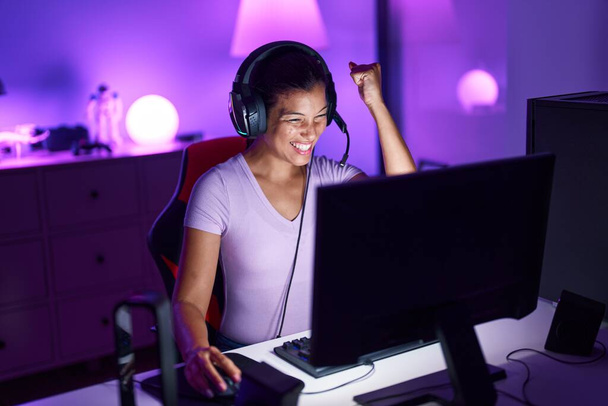 Mladá krásná hispánská žena streamer hrát videohru s vítězným výrazem v herně - Fotografie, Obrázek