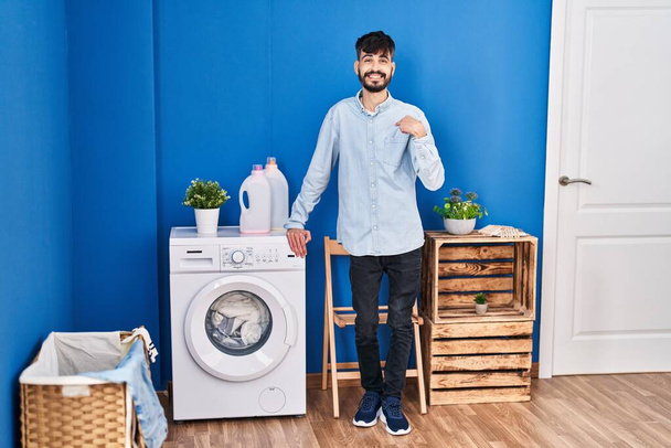 Joven hombre hispano con barba haciendo la colada de pie en la lavandería señalando con el dedo a uno mismo sonriendo feliz y orgulloso  - Foto, Imagen