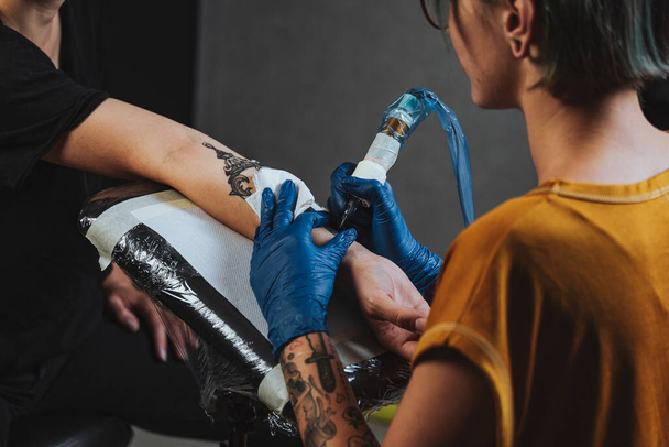 Close up tattoo artist demonstrates the process of getting black tattoo with paint. Master works in blue sterile gloves. Master of tattoo fill circuit tattoo. - Photo, Image