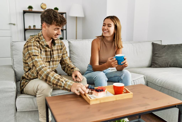 Fiatal spanyol pár mosolyog, boldogan reggeliznek otthon a kanapén.. - Fotó, kép