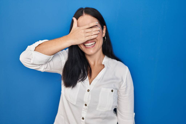 Jonge Latijns-Amerikaanse vrouw die over een blauwe achtergrond staat te glimlachen en met de hand op het gezicht ogen bedekt voor verrassing. blind concept.  - Foto, afbeelding