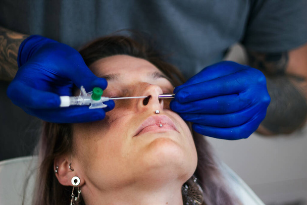 Portrait of a woman getting her nose pierced. Man showing a process of piercing nose with steril neadle and latex gloves. Nostril Piercing Procedure - Photo, Image