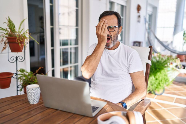 Middelbare leeftijd man met behulp van computer laptop thuis geeuwen moe bedekken half gezicht, oog en mond met de hand. gezicht doet pijn in pijn.  - Foto, afbeelding