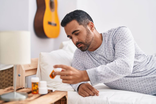Young hispanic man lying on bed holding pills at bedroom - Photo, Image