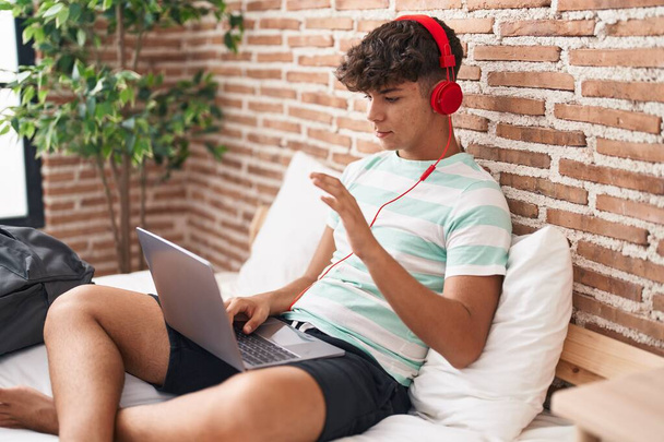 Adolescent hispanique utilisant un ordinateur portable à la maison assis sur la chambre à coucher regardant positif et heureux debout et souriant avec un sourire confiant montrant les dents  - Photo, image
