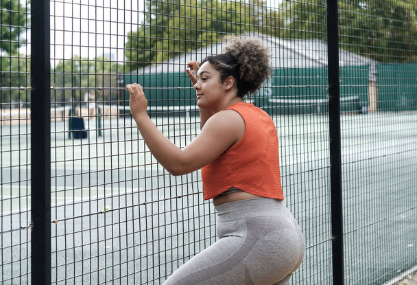 Μια νεαρή αθλήτρια κάνει τρίψιμο στο γήπεδο του τένις και κοιτάζει μέσα με αποφασιστικότητα. Σγουρά μαλλιά, πορτοκαλί αμάνικο μπλουζάκι και γκρι κολάν. Υπέρβαση έννοιας. - Φωτογραφία, εικόνα
