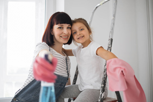 Beautiful young woman and her little daughter are smiling while preparing cleaning window at home using detergent and rags - Foto, imagen