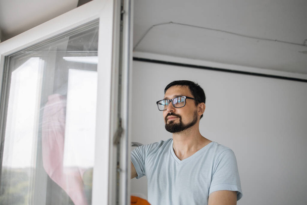 Close up portrait of bearded caucasian man in eyeglasses, sitting on stepladder holding sponge for washing windows and detergents, cleaning the apartment after renovation is completed. - Fotoğraf, Görsel