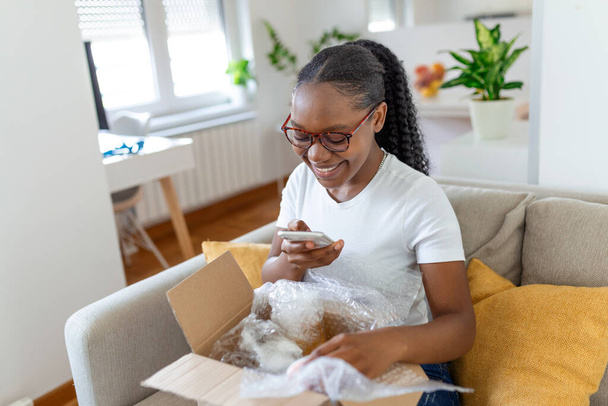 Joven satisfecha feliz mujer africana mujer shopaholic cliente sentarse en sofá desempaquetar paquete caja de entrega, concepto de envío de compras en línea. tomar fotos del producto para publicar en las redes sociales - Foto, imagen