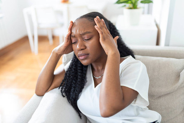Portrait of a young black girl sitting on the couch at home with a headache and pain. Beautiful woman suffering from chronic daily headaches. Sad woman holding her head because sinus pain - Photo, Image