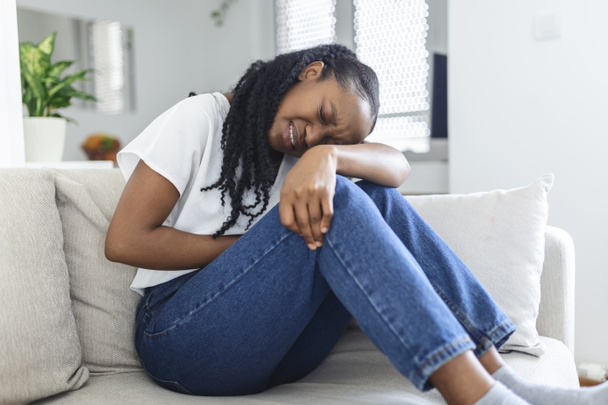 African American Woman in painful expression holding hands against belly suffering menstrual period pain, lying sad on home bed, having tummy cramp in female health concept - 写真・画像