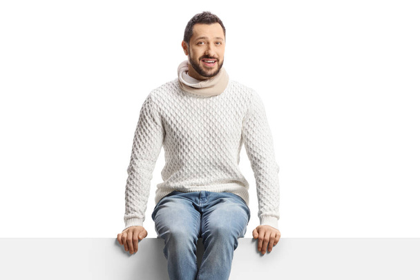 Casual young man in a woolen jumper and sitting on a blank panel isolated on white background - Foto, imagen