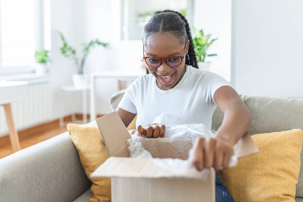 Glückliche junge Frau sitzt auf der Couch im Wohnzimmer und packt Pappschachtel aus, kauft Waren im Internet, lächelt aufgeregt Millennialmädchen öffnet Pappkarton, bestellt online, gutes Lieferkonzept - Foto, Bild