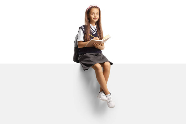Schoolgirl sitting on a blank panel and writing a homework isolated on white background - 写真・画像