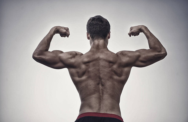 Athlete with bare torso on grey sky. Bodybuilder showing muscles, biceps and triceps. Man or sportsman flexing arms with fists, back view. Fitness and sport. Healthy lifestyle concept - Photo, Image