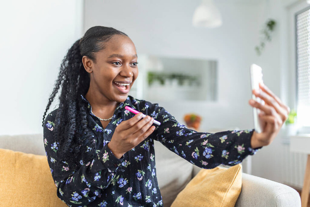 Feliz, mujer africana embarazada está mostrando su prueba de embarazo y tomando selfie hacer videollamada. Mujer feliz tomando fotos de la prueba de embarazo con el teléfono móvil y la publicación de la imagen en las redes sociales. - Foto, Imagen