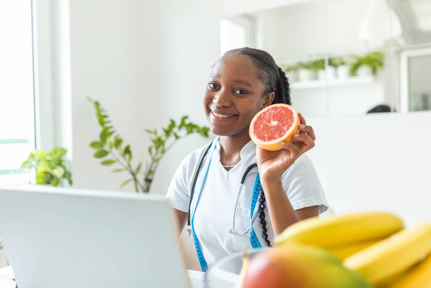 Portrait of young smiling female nutritionist in the consultation room. Nutritionist desk with healthy fruit, juice and measuring tape. Dietitian working on diet plan. - Photo, Image