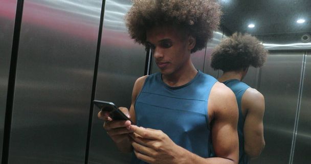Молодой афроамериканец проверяет мобильное устройство в лифте, человек выходит из лифта - Фото, изображение