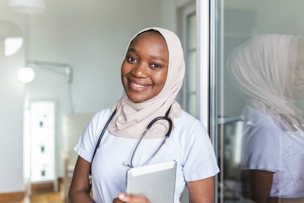 Giovane donna musllim medico clinico africano in scrub utilizzando touchpad durante la comunicazione con i pazienti online - Foto, immagini