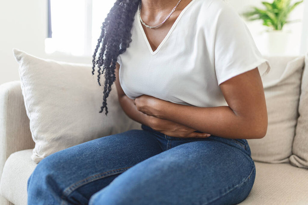 African American Woman in painful expression holding hands against belly suffering menstrual period pain, lying sad on home bed, having tummy cramp in female health concept - Fotografie, Obrázek