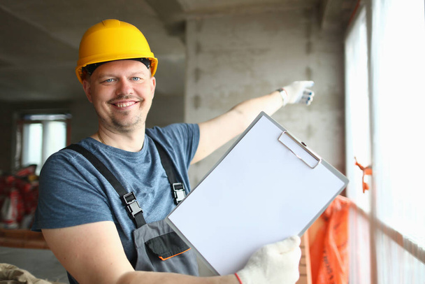 Trabajador constructor sonriente en sombrero duro sosteniendo hoja de papel en blanco en el portapapeles. Concepto de obras y servicios de renovación de apartamentos - Foto, imagen
