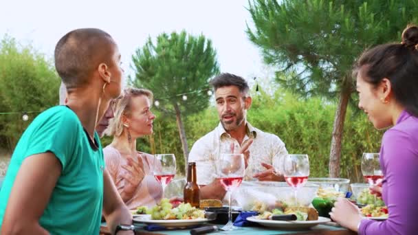 houkutteleva aikuinen latinomies puhuu ystäviensä kanssa, kun taas kaksi ystävää paahtaa hänen viinilasit takapihalla illalliskutsuilla. Laadukas 4k hidastettuna kuvamateriaalia - Materiaali, video