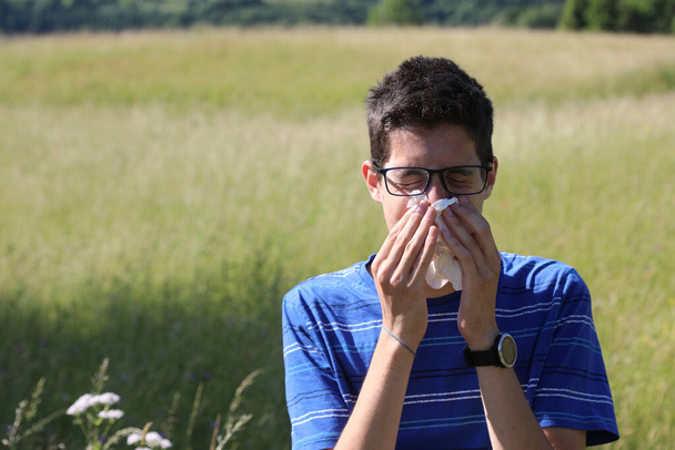 giovane ragazzo con gli occhiali soffiando il naso a causa di allergia nel prato in primavera - Foto, immagini