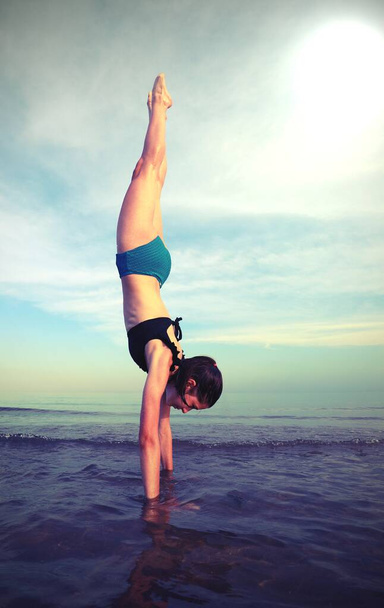 Kız tamamen dikey olarak jimnastik yapıyor. Kafası aşağıda ve bacakları deniz kıyısında eski tonlu efektlerle. - Fotoğraf, Görsel