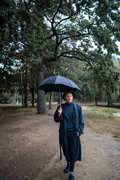 young english man in stylish vintage coat and black hat walking in rainy forest - Foto, afbeelding