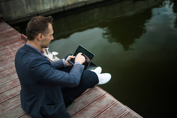 Alegre y exitoso hombre de negocios trabaja al aire libre en puente de madera cerca del lago tiene una tableta - Foto, imagen