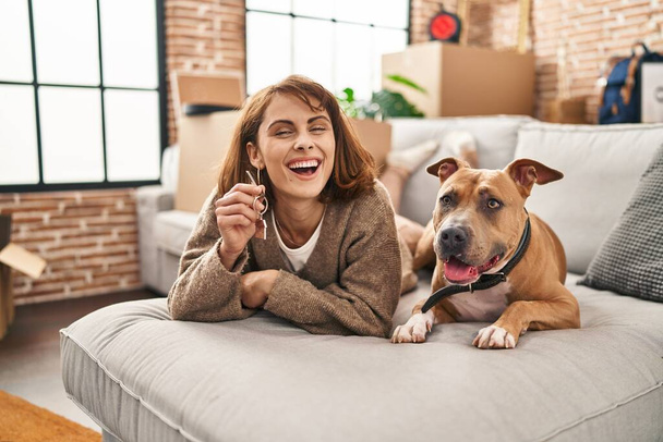 Νεαρή καυκάσια γυναίκα κρατώντας το κλειδί του νέου σπιτιού που βρίσκεται στον καναπέ με το σκυλί στο νέο σπίτι - Φωτογραφία, εικόνα