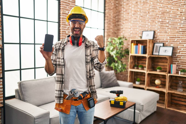 Junger hispanischer Mann mit Bart arbeitet bei Renovierungsarbeiten zu Hause und hält stolz sein Smartphone in der Hand und feiert Sieg und Erfolg mit erhobenen Armen  - Foto, Bild