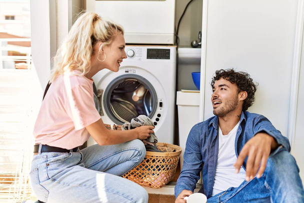 Young couple smiling happy drinking coffee while doing laundry at home. - Foto, afbeelding