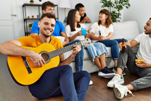 Ομάδα νεαρών φίλων στο πάρτι χαμογελώντας ευτυχισμένοι και παίζοντας κλασική κιθάρα στο σπίτι. - Φωτογραφία, εικόνα