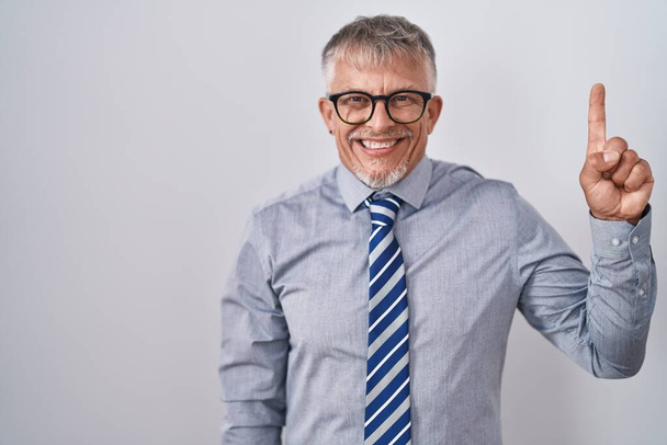 Hispanic business man with grey hair wearing glasses showing and pointing up with finger number one while smiling confident and happy.  - Photo, image