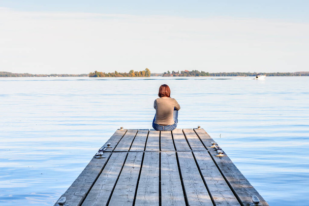 Μοναχική γυναίκα που κάθεται στην άκρη μιας ξύλινης προβλήτας στην όχθη ενός μεγάλου ποταμού μια ηλιόλουστη φθινοπωρινή μέρα. Saint Lawrence river, Thousand Islands National Park, ON, Καναδάς. - Φωτογραφία, εικόνα