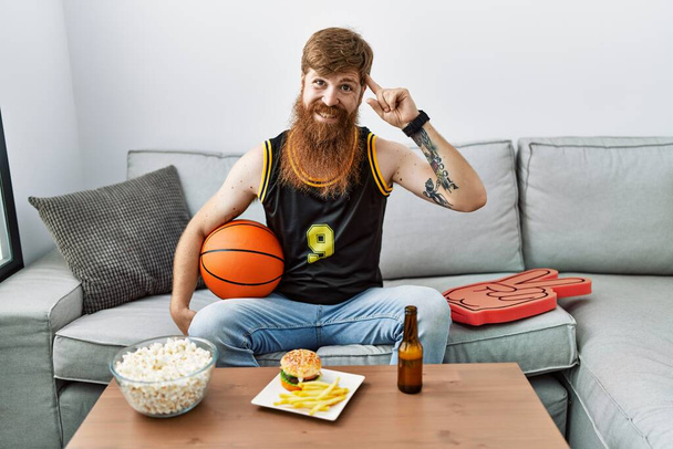Kaukázusi férfi hosszú szakállal gazdaság kosárlabda éljenzés tv játék mosolygós mutató fej egy ujj, nagy ötlet vagy gondolat, jó memória  - Fotó, kép