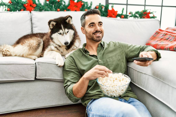 Jonge Spaanse man kijken film zitten op de bank met hond door kerst decor thuis - Foto, afbeelding