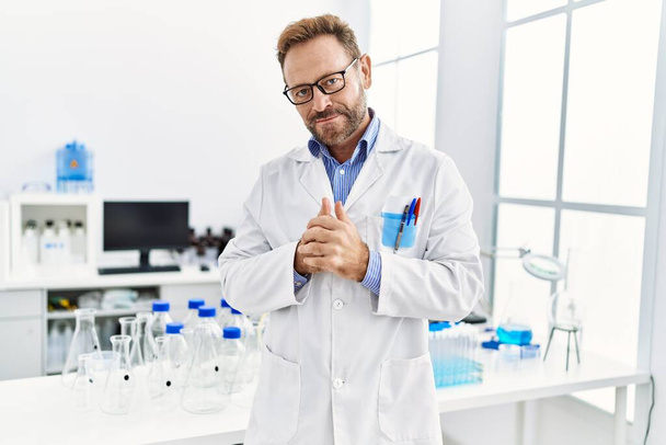 Hombre hispano de mediana edad vistiendo uniforme científico parado en el laboratorio - Foto, Imagen