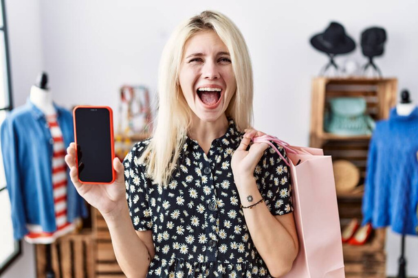 Młoda blondynka trzyma torby na zakupy pokazując ekran smartfona świętując szalony i zdumiony sukcesem z otwartymi oczami krzycząc podekscytowany.  - Zdjęcie, obraz