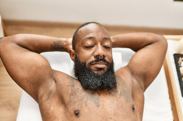 Giovane uomo afroamericano rilassato con le mani sulla testa sdraiato sul lettino da massaggio al centro estetico - Foto, immagini