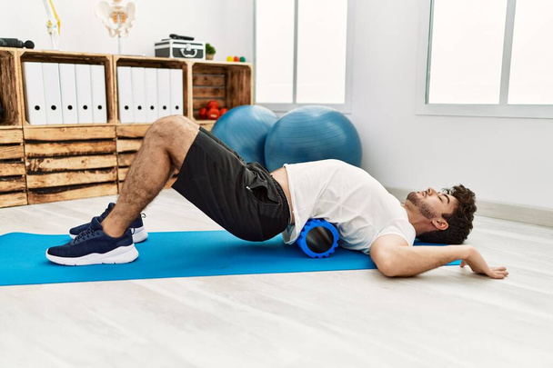 Young hispanic man doing rehab using foam roller at clinic - Foto, afbeelding