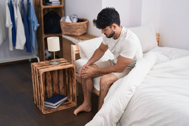 Young hispanic man suffering for knee injury sitting on bed at bedroom - Photo, image