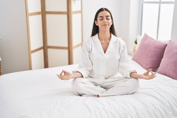 Young hispanic woman doing yoga exercise sitting on bed at bedroom - Photo, image