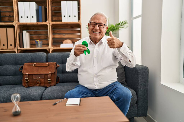 Thérapeute senior avec des cheveux gris tenant le ruban vert de soutien pour la sensibilisation à la santé mentale souriant heureux et positif, pouce levé faire excellent et signe d'approbation  - Photo, image