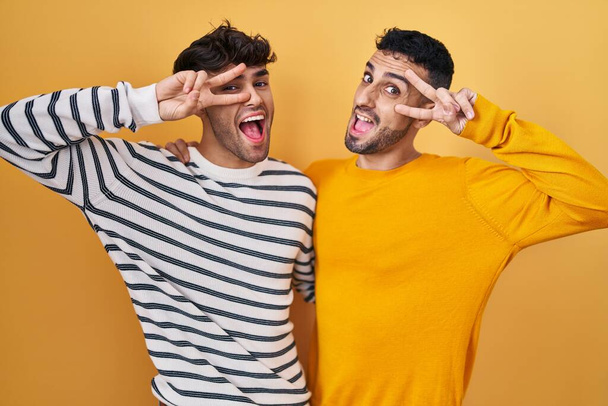 Joven pareja gay hispana de pie sobre fondo amarillo haciendo símbolo de paz con los dedos sobre la cara, sonriendo alegre mostrando la victoria  - Foto, Imagen